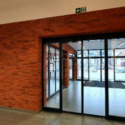 Wnętrze budynku z wykończoną ścianą z czerwonej cegły i automatycznymi drzwiami przesuwnymi z czarnymi ramami, widok na zewnątrz z placem budowy.