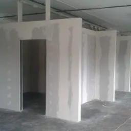 Wnętrze z surowymi ścianami działowymi z płyt gipsowo-kartonowych, widoczne miejsca łączeń płyt wymagające szpachlowania, surowy betonowy podkład podłogowy, widoczne instalacje pod sufitem.