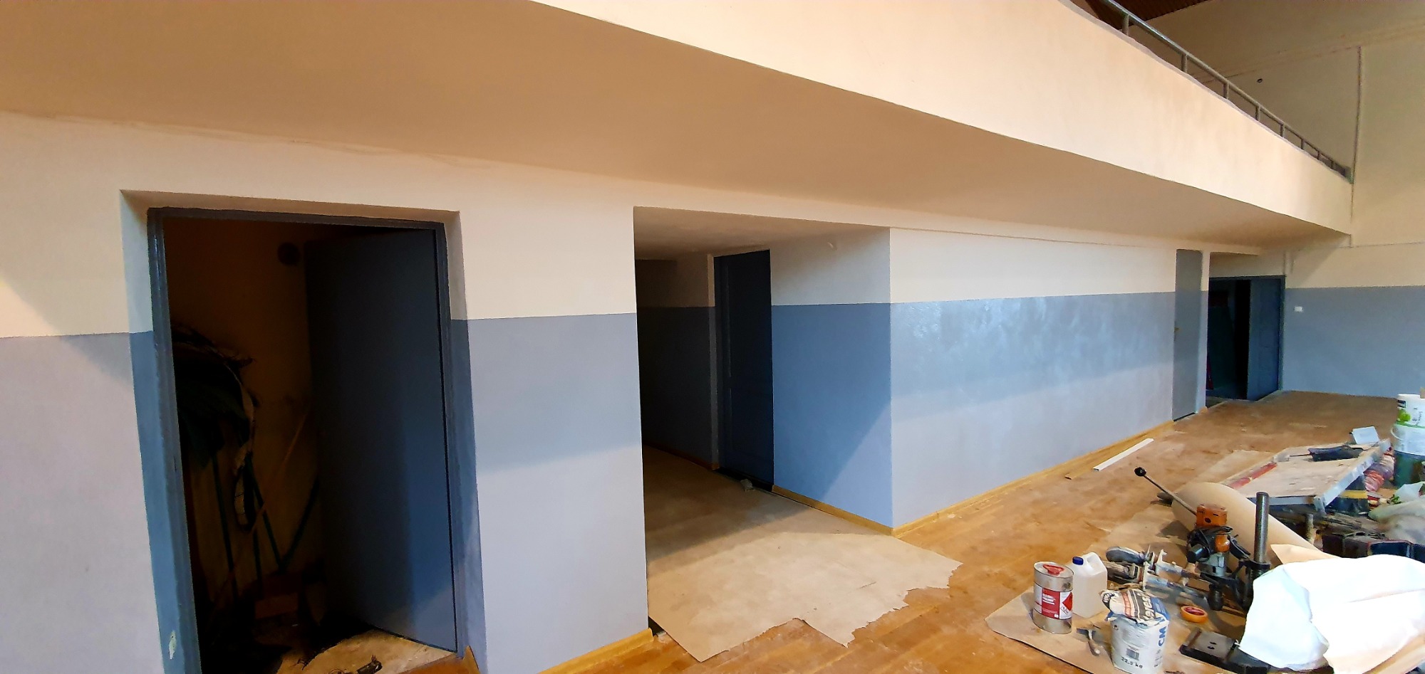 Wnętrze budynku w trakcie malowania, ściany pomalowane na dwa kolory - biały u góry i szary na dole, widoczne narzędzia malarskie i materiały budowlane, podłoga zabezpieczona kartonem.