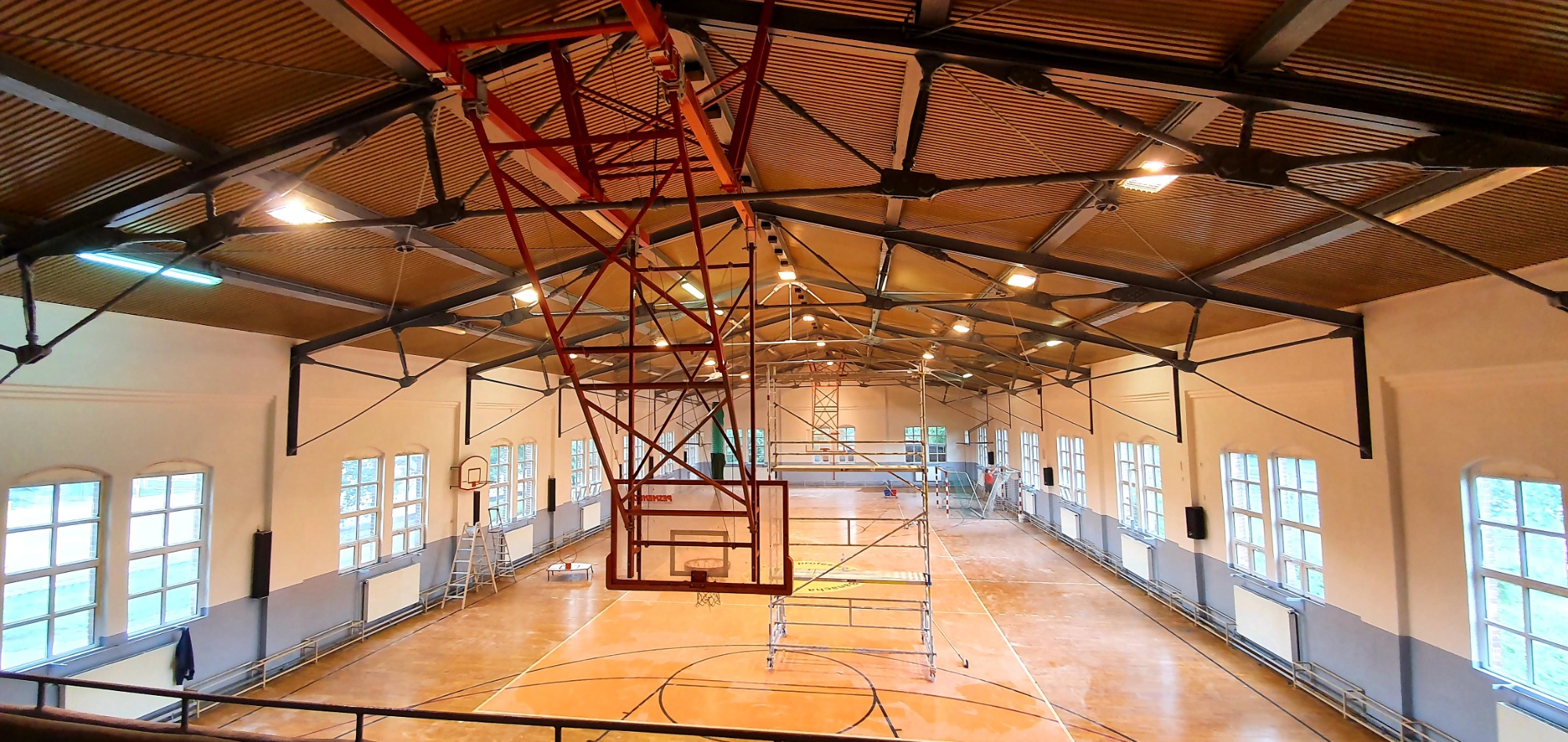 Wnętrze hali sportowej z widocznym rusztowaniem i pracami na wysokości, oświetlenie jarzeniowe i naturalne z okien, metalowa konstrukcja dachu.
