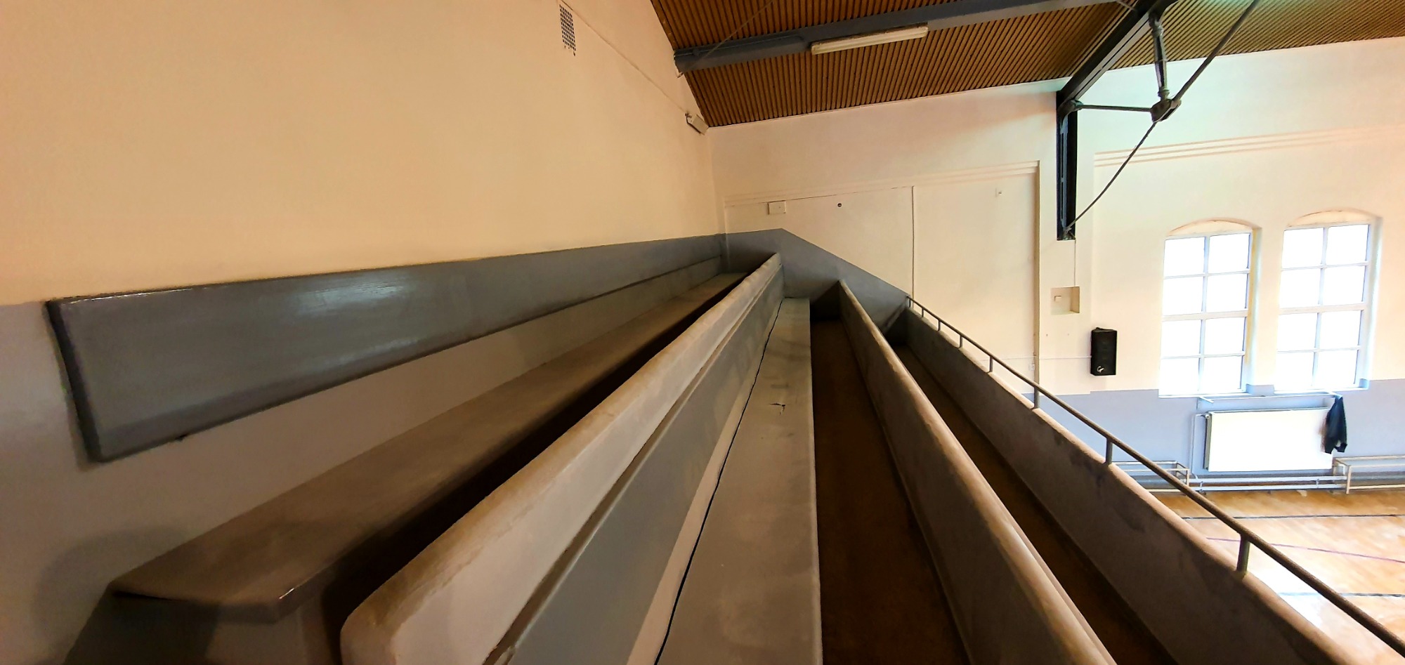 Widok z góry na odnowione, szare i kremowe ściany hali sportowej z drewnianym parkietem, oświetlone przez okna z białymi ramami, z widocznymi elementami konstrukcyjnymi pod sufitem.