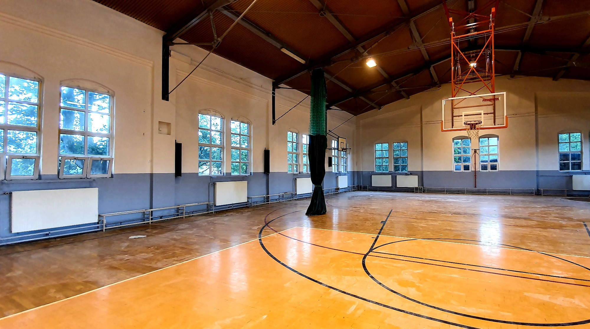 Wnętrze przestronnej sali gimnastycznej z parkietem w odcieniach brązu i żółci, widoczne linie boiska, kosz do koszykówki na ścianie, wysokie okna z widokiem na drzewa oraz grzejniki pod oknami.
