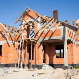 Surowy stan domu jednorodzinnego z czerwonej cegły z drewnianą konstrukcją dachu, widoczne rusztowania i fundamenty z izolacją, na tle błękitnego nieba.