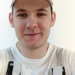 Portret uśmiechniętego mężczyzny w białej koszulce i roboczych szelkach, z białym tłem ściany.