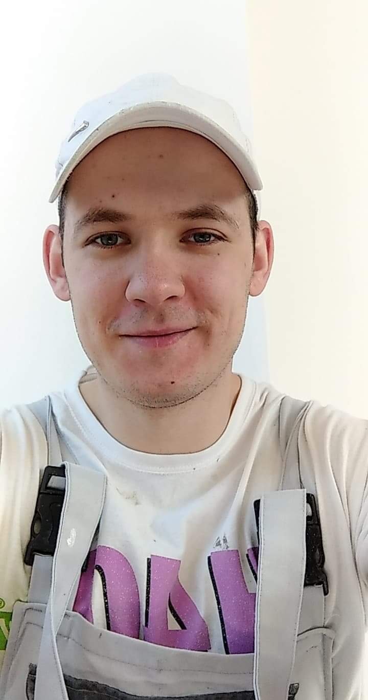 Portret uśmiechniętego mężczyzny w białej koszulce i roboczych szelkach, z białym tłem ściany.