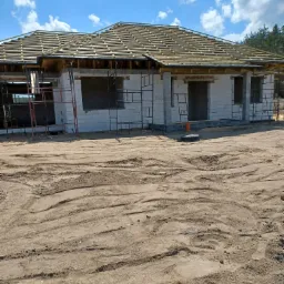 Niedokończony dom jednorodzinny z białych bloczków, z drewnianą konstrukcją dachu, otoczony piaskiem i rusztowaniami, w słoneczny dzień.