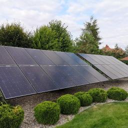 Mako Maciej Marcinów - Dwa rzędy paneli słonecznych ustawione na żwirowym podłożu, otoczone kulistymi krzewami i trawnikiem, z budynkiem w tle.