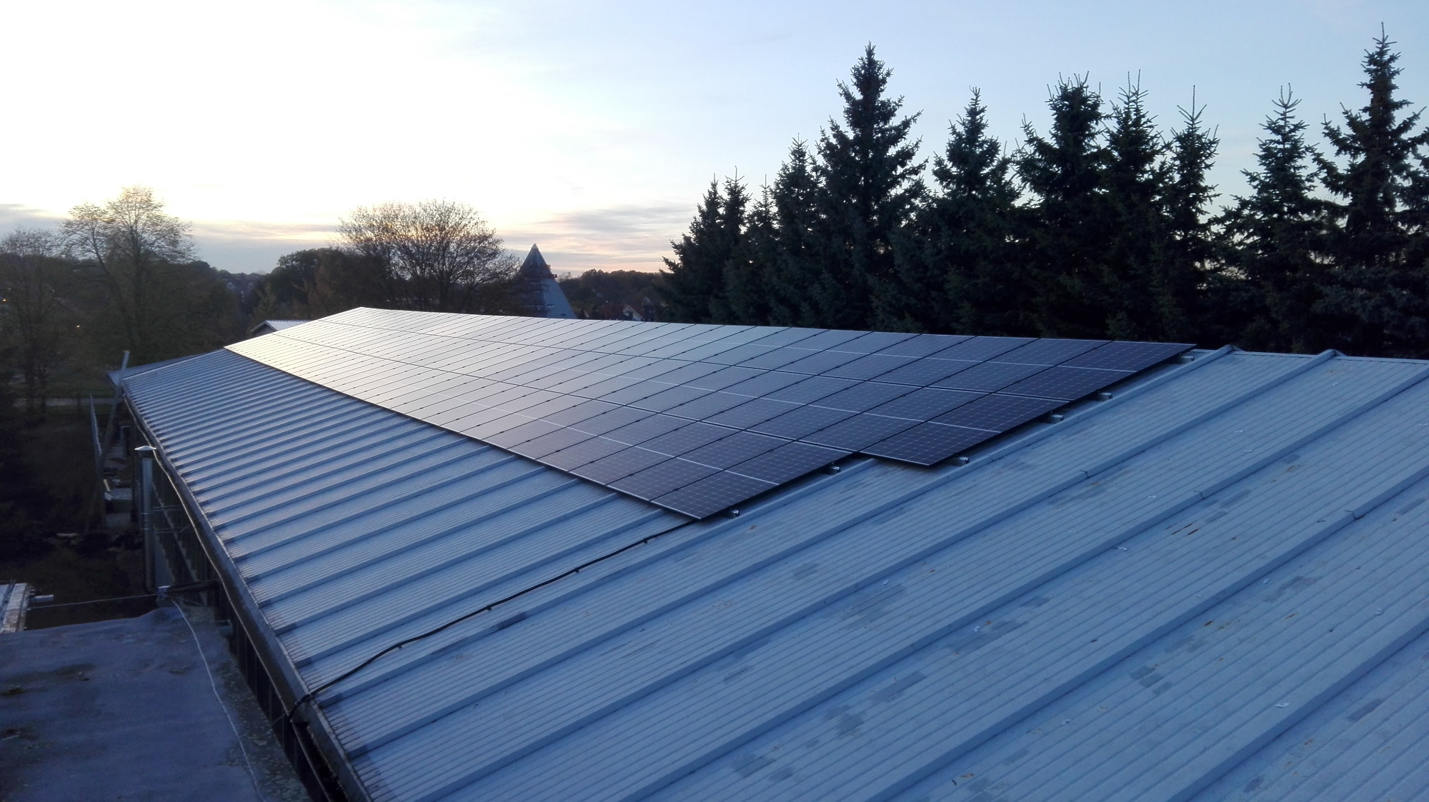 Panele fotowoltaiczne zamontowane na metalowym dachu o trapezowym profilu, widok z góry, w tle drzewa iglaste i fragmenty zabudowy miejskiej w oddali o zmierzchu.