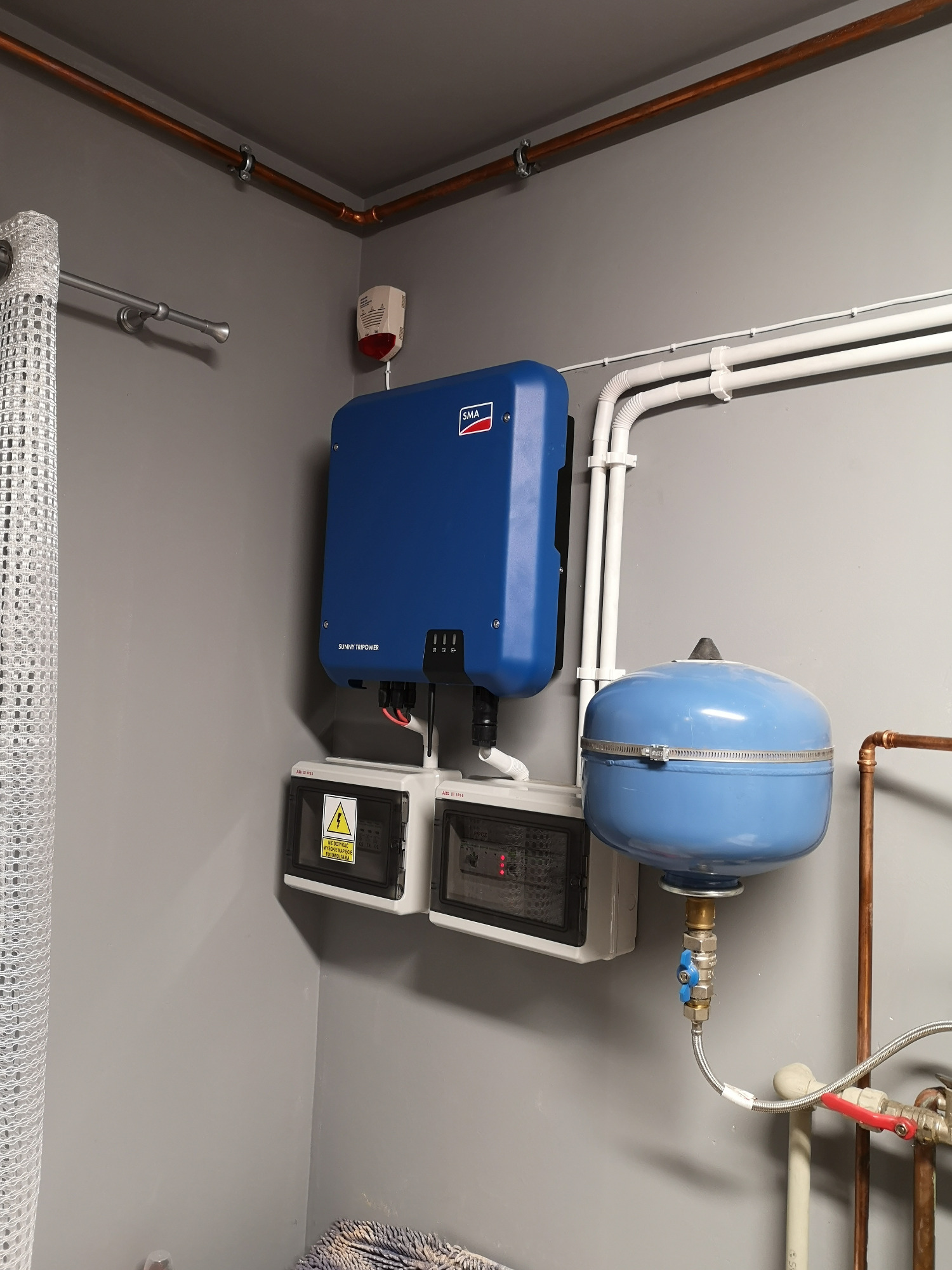 Niebieski inwerter SMA Sunny Tripower zamontowany na szarej ścianie, obok białe skrzynki z bezpiecznikami i niebieski zbiornik ciśnieniowy, widoczne miedziane rury instalacji.