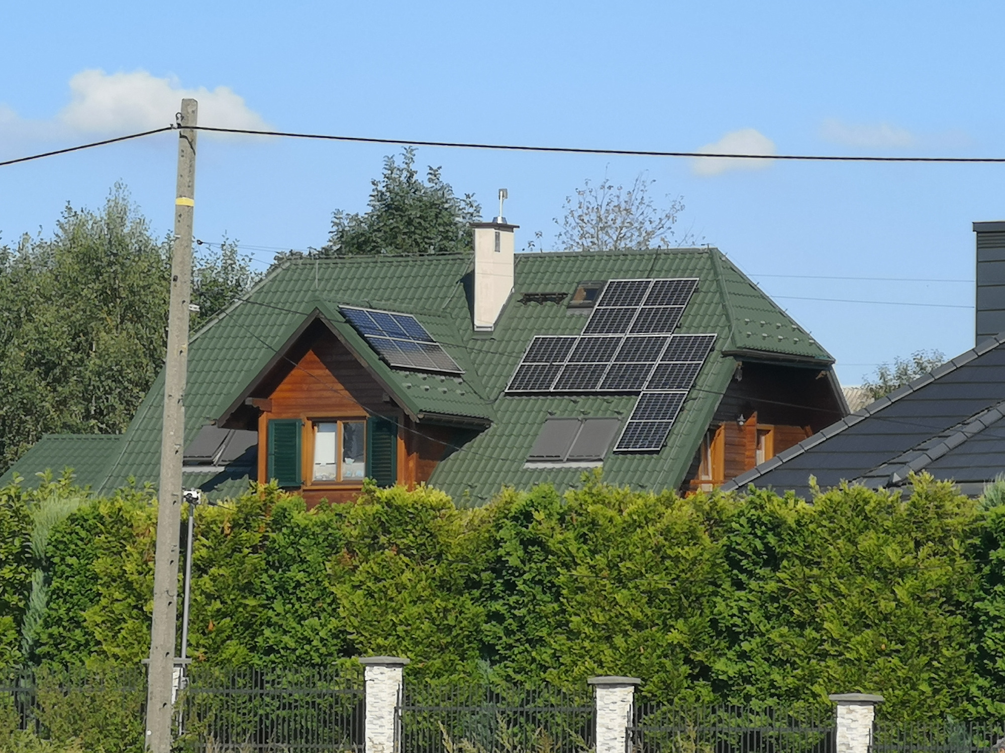 Dom z zielonym dachem i panelami słonecznymi, częściowo zasłonięty żywopłotem, widoczny słup elektryczny po lewej stronie.