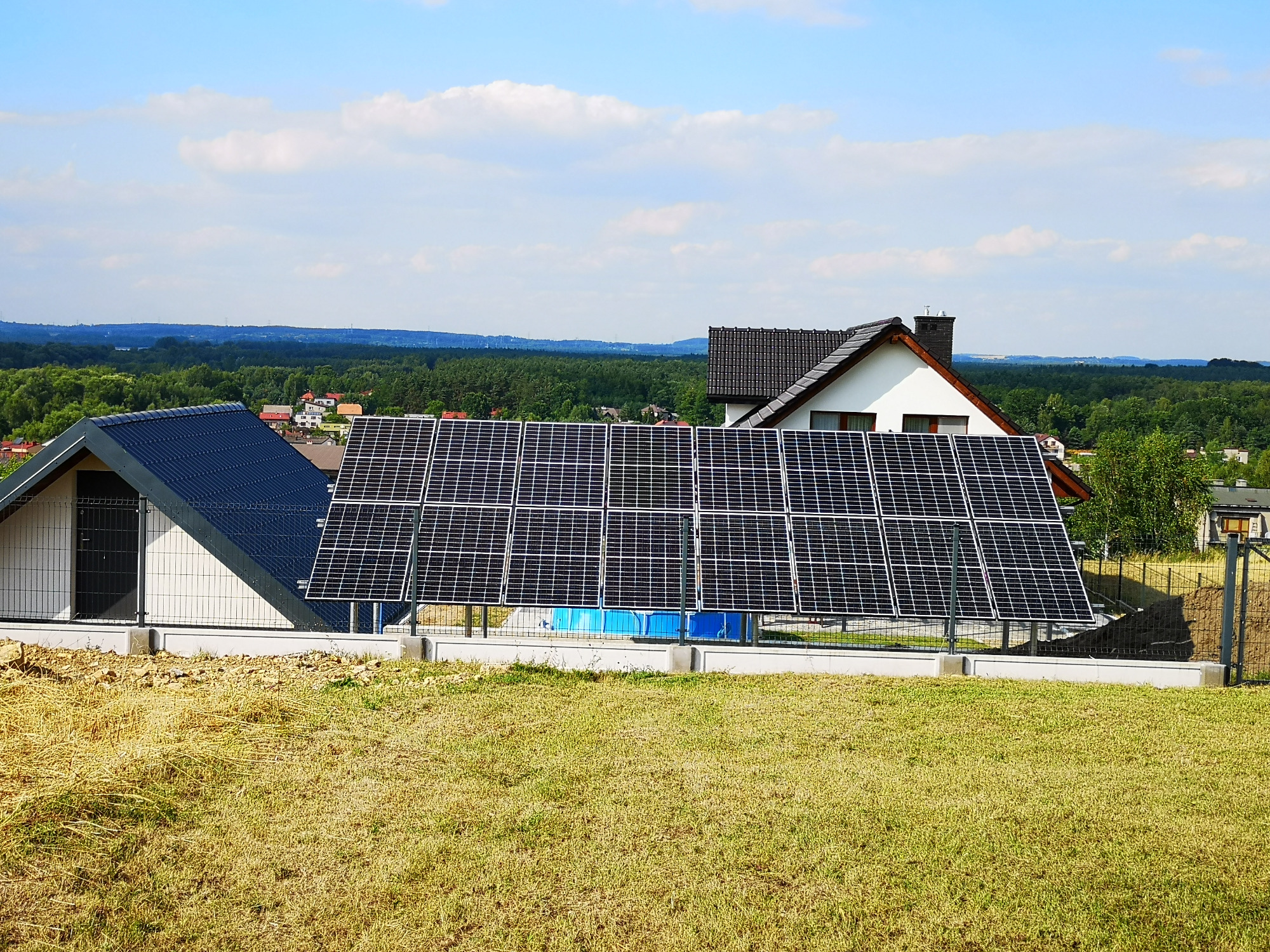 Rząd paneli słonecznych na metalowej konstrukcji w ogrodzie, z domami w tle i widokiem na zalesione wzgórza pod błękitnym niebem z chmurami.