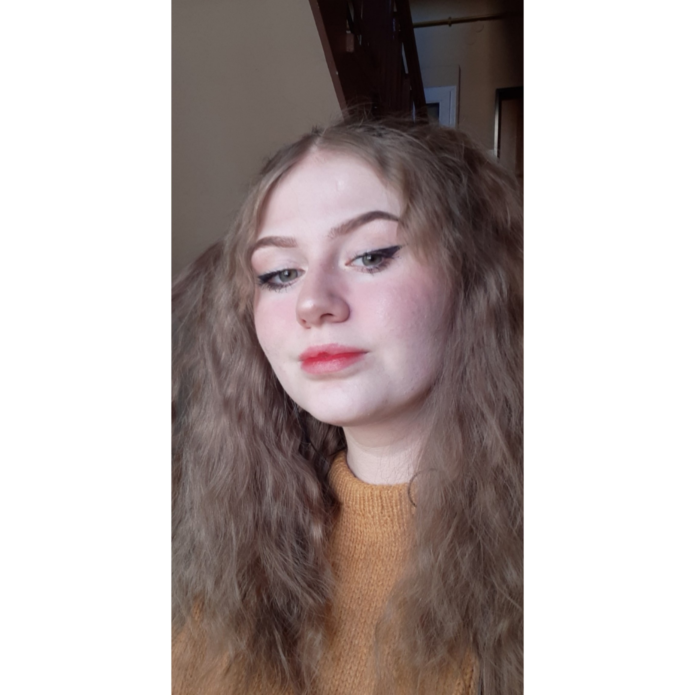 Portret młodej kobiety z blond włosami spiętymi w dwa kucyki, z delikatnym makijażem i w musztardowym swetrze, na tle fragmentu schodów.