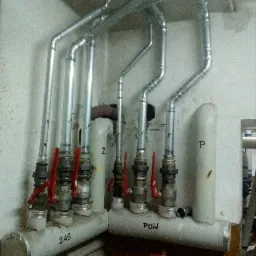 Rozdzielacze z zaworami i rurami w piwnicy, oznaczone napisem 'ZAS' i 'POW' na szarym tle ściany, widoczne elementy instalacji centralnego ogrzewania.