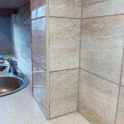 Fragment wykończonej kuchni z beżowymi płytkami imitującymi kamień, widoczny zlew i blat z drobnymi plamkami, oraz gniazdko elektryczne na ścianie.