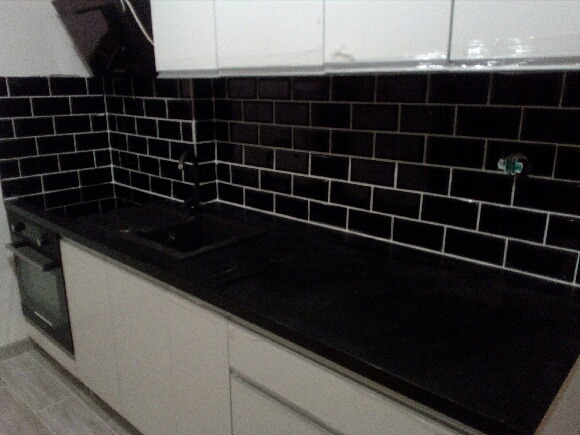 Nowoczesna kuchnia z czarnymi, błyszczącymi płytkami ułożonymi w cegiełkę, białe szafki dolne i górne, czarny blat i zlewozmywak, widoczny fragment kuchenki.