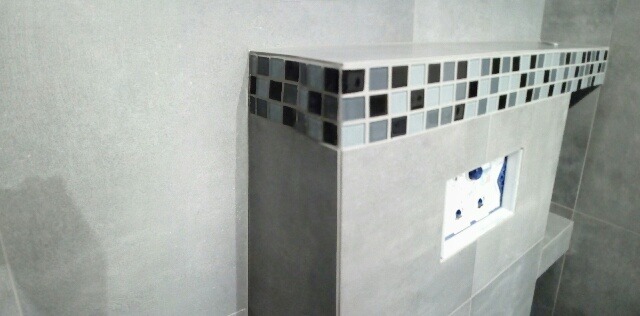 Obudowa stelaża WC wykończona płytkami ceramicznymi w odcieniach szarości i czerni, z widocznym otworem na przycisk spłukujący.