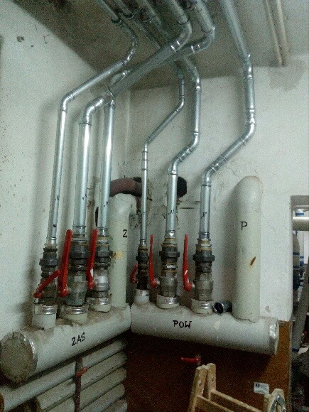 Rozdzielacze z zaworami i rurami w piwnicy, oznaczone napisem 'ZAS' i 'POW' na szarym tle ściany, widoczne elementy instalacji centralnego ogrzewania.