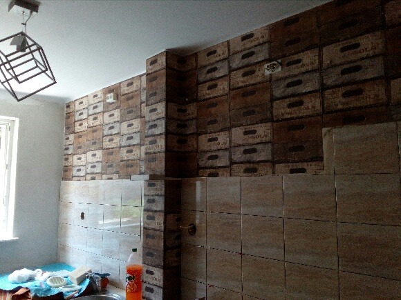 Ściana wyłożona płytkami imitującymi kamień, górna część pokryta tapetą z motywem skrzynek, widoczny fragment oświetlenia sufitowego, butelka napoju i fragment zlewu.