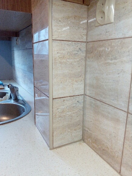 Fragment wykończonej kuchni z beżowymi płytkami imitującymi kamień, widoczny zlew i blat z drobnymi plamkami, oraz gniazdko elektryczne na ścianie.