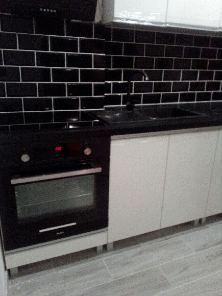 Nowoczesna kuchnia z czarnymi, błyszczącymi płytkami na ścianie, białymi szafkami i czarnym piekarnikiem Amica.