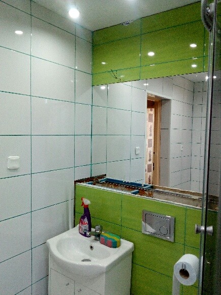 Łazienka w trakcie remontu: białe i zielone płytki, niedokończona zabudowa lustra, umywalka z płynem do czyszczenia, widoczne elementy instalacji.
