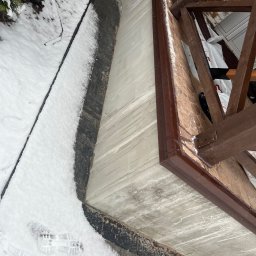 Chrzan-Dach - Krawędź dachu z brązowym obramowaniem, pokryta osadzonym śniegiem, widoczna z perspektywy od dołu, z fragmentem drewnianej konstrukcji dachu, częściowo ośnieżonym chodnikiem i odciskiem buta w śniegu.