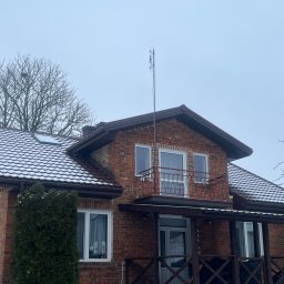 Chrzan-Dach - Dwukondygnacyjny dom z czerwonej cegły z brązowym dachem pokrytym lekkim śniegiem, widoczne rynny i wysoka antena na dachu.