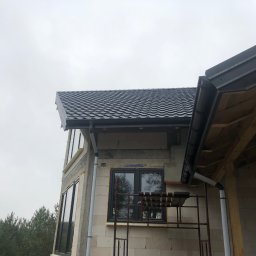 Chrzan-Dach - Fragment domu w trakcie budowy z zamontowanymi oknami, czarną blachodachówką na dachu i ciemnymi rynnami. Widoczny fragment rusztowania i las w tle.