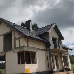 Chrzan-Dach - Dom w stanie surowym otwartym z zamontowanym dachem z ciemnej dachówki i rynnami, widoczne obróbki blacharskie. W tle pochmurne niebo.