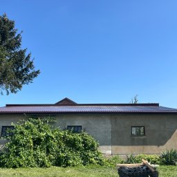 Chrzan-Dach - Budynek gospodarczy z nowym, ciemnobrązowym dachem z blachy trapezowej, częściowo zasłonięty bujną zielenią, na trawniku stos ułożonych arkuszy blachy.