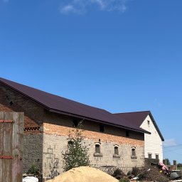 Chrzan-Dach - Budynek gospodarczy z ciemnobrązowym dachem z blachy, widok na fragment elewacji z cegły, okna z łukami i stertę piasku na pierwszym planie.
