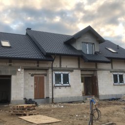 Chrzan-Dach - Dom w stanie surowym zamkniętym z ciemną dachówką i rynnami, widoczne okna dachowe i orynnowanie, teren wokół w trakcie prac budowlanych, pracownik w garażu.