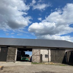 Chrzan-Dach - Duży, murowany budynek gospodarczy z nowym, ciemnoszarym dachem z blachy trapezowej, otwartymi drewnianymi wrotami i przyczepą w środku, na tle błękitnego nieba z białymi chmurami.