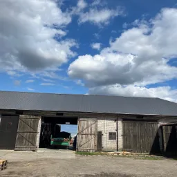 Duży, murowany budynek gospodarczy z nowym, ciemnoszarym dachem z blachy trapezowej, otwartymi drewnianymi wrotami i przyczepą w środku, na tle błękitnego nieba z białymi chmurami.