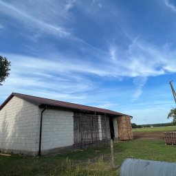 Chrzan-Dach - Budynek gospodarczy z szarej cegły z brązowym dachem i rynnami, częściowo otwarte drewniane wrota, w tle błękitne niebo z chmurami, na palecie ułożone dachówki.
