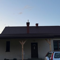Chrzan-Dach - Dom jednorodzinny z ciemnobrązowym dachem z dachówki, widoczny komin z cegły i fragment srebrnego samochodu. Ujęcie z dołu, w tle błękitne niebo.