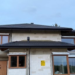 Chrzan-Dach - Dom w trakcie budowy z czarnym dachem z dachówki ceramicznej, widoczne okna z drewnianą stolarką i fragment niezakończonego krycia dachu po lewej stronie.