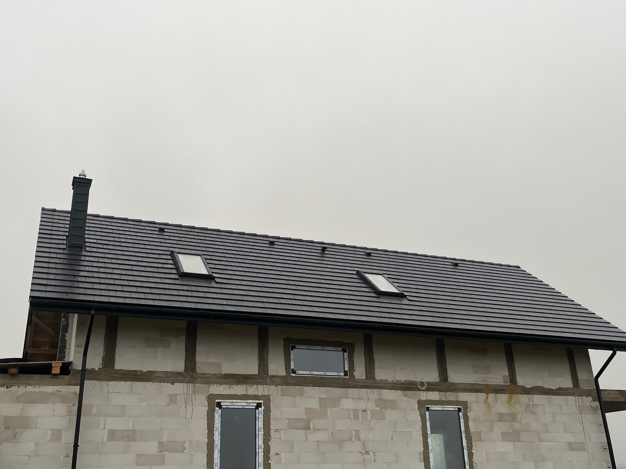 Nowy dach z czarnej dachówki ceramicznej, z oknami dachowymi i kominem, na budynku w trakcie budowy z białego bloczka. Widoczne rynny i obróbki blacharskie.