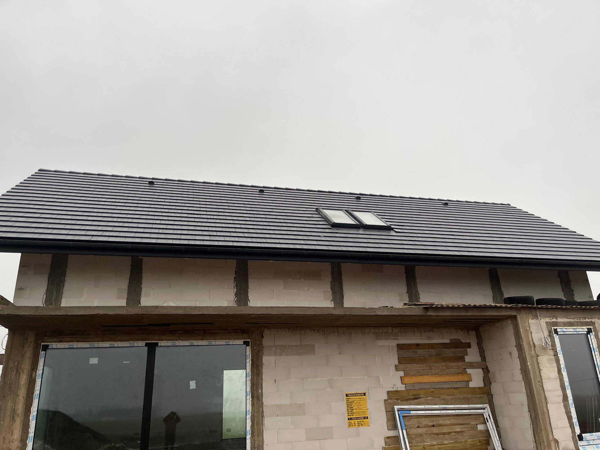 Nowy dach z dachówką ceramiczną w kolorze grafitowym, z dwoma oknami dachowymi, na surowym budynku w trakcie budowy, widok z dołu na tle pochmurnego nieba.