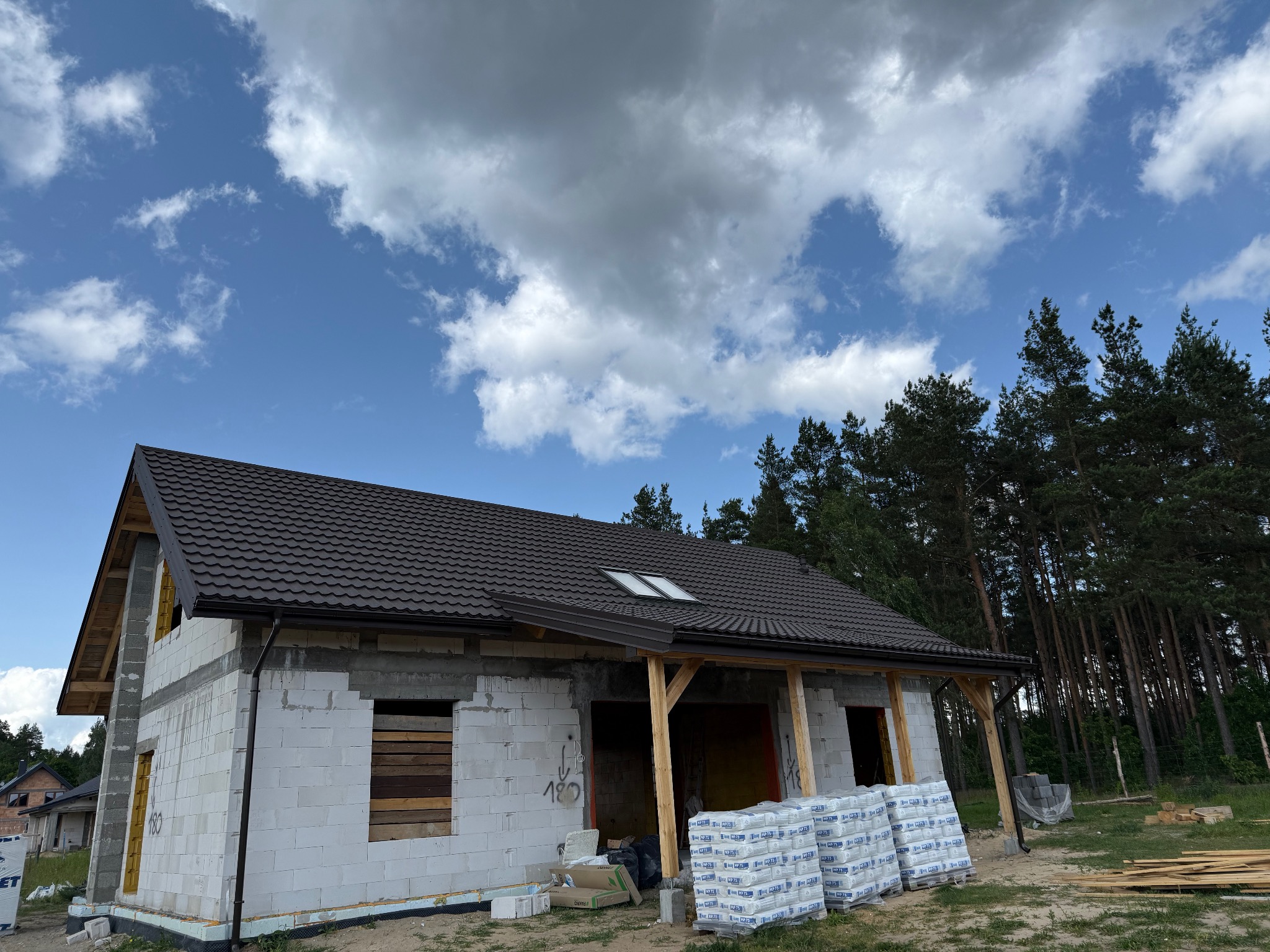 Dom w trakcie budowy z brązowym dachem i rynnami, widoczne materiały budowlane. Tło stanowią drzewa i błękitne niebo z chmurami.