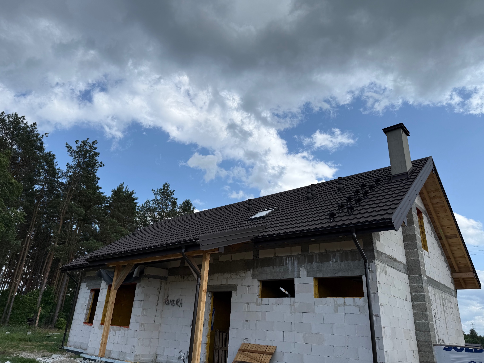 Dom w trakcie budowy z ciemnym dachem i rynnami, widoczne bloczki, drewniane elementy konstrukcyjne i fragment lasu w tle pod pochmurnym niebem.