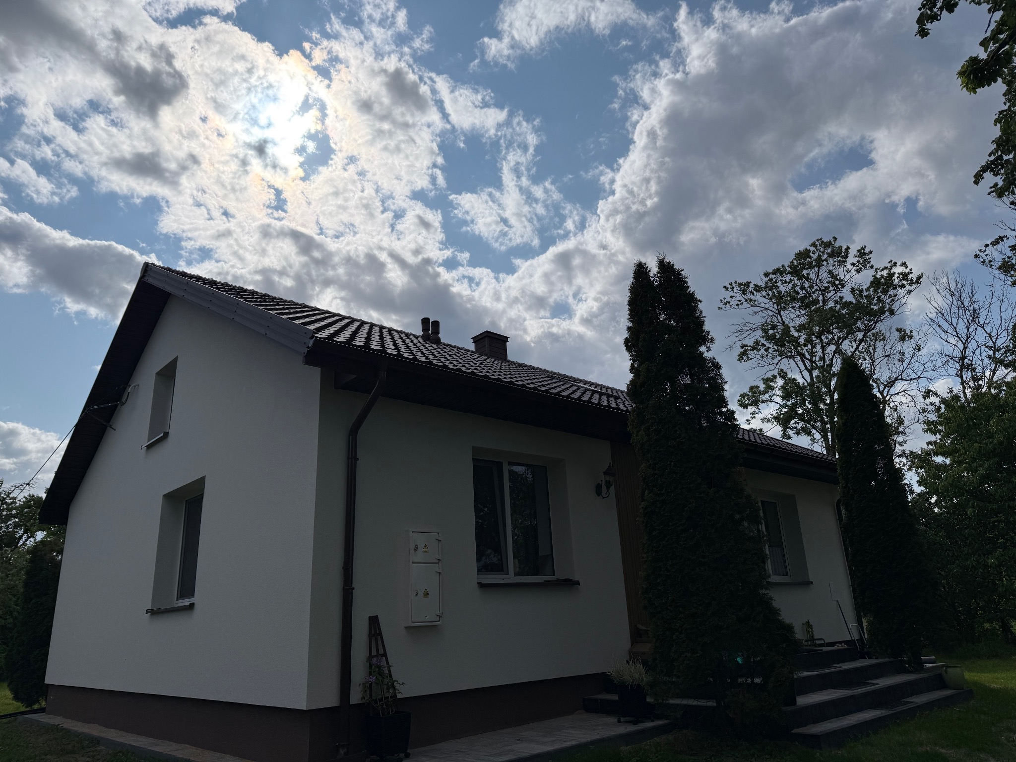 Dom jednorodzinny z ciemną dachówką w słoneczny dzień. Widoczne obróbki blacharskie i rynny. Tło stanowią drzewa i błękitne niebo z chmurami.