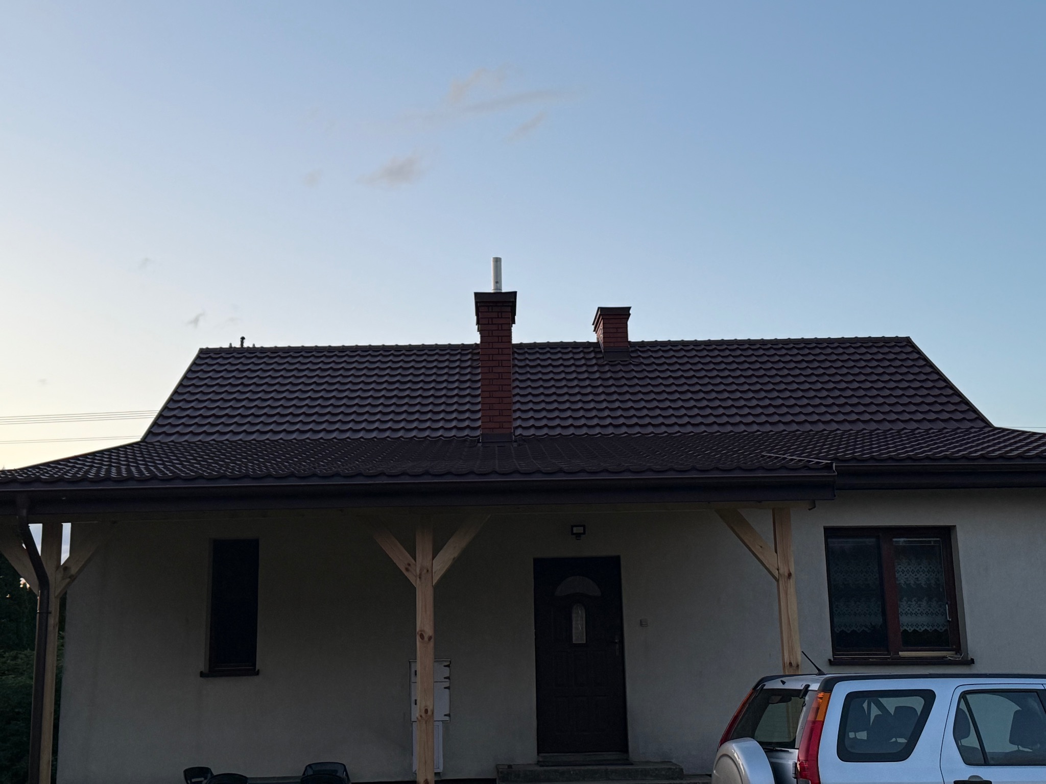 Dom jednorodzinny z ciemnobrązowym dachem z dachówki, widoczny komin z cegły i fragment srebrnego samochodu. Ujęcie z dołu, w tle błękitne niebo.