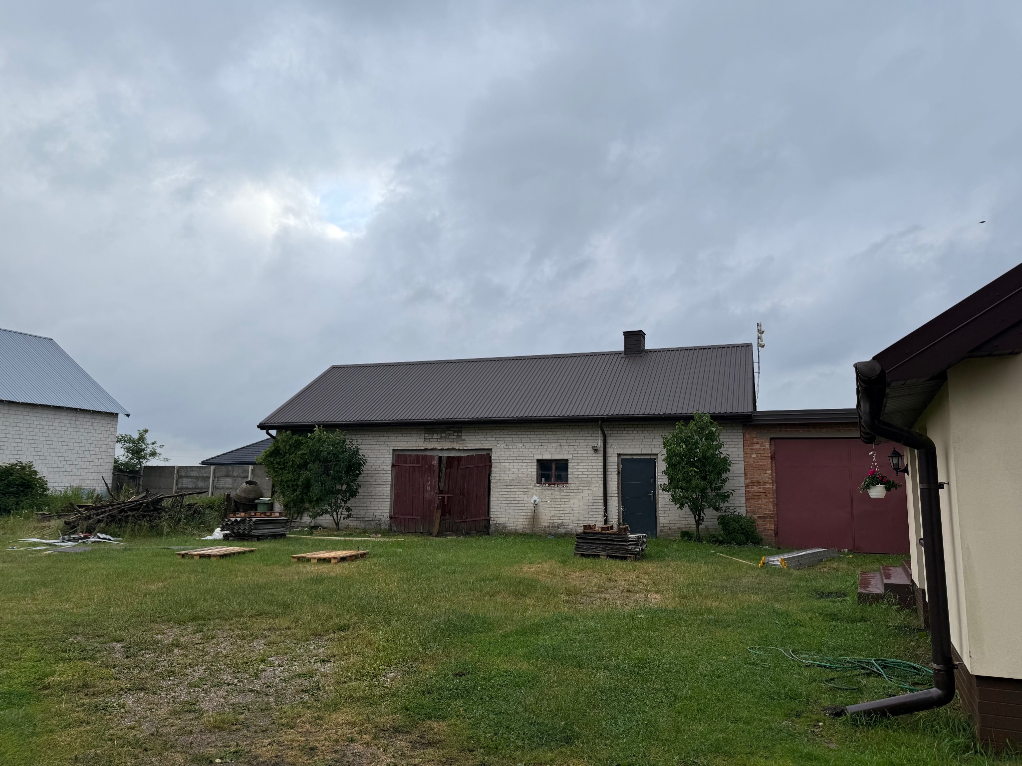 Budynek gospodarczy z brązowym dachem z blachy falistej, widoczny z trawnika w pochmurny dzień. Na dachu komin. Częściowo widoczne rynny. Na trawniku palety i drabina.
