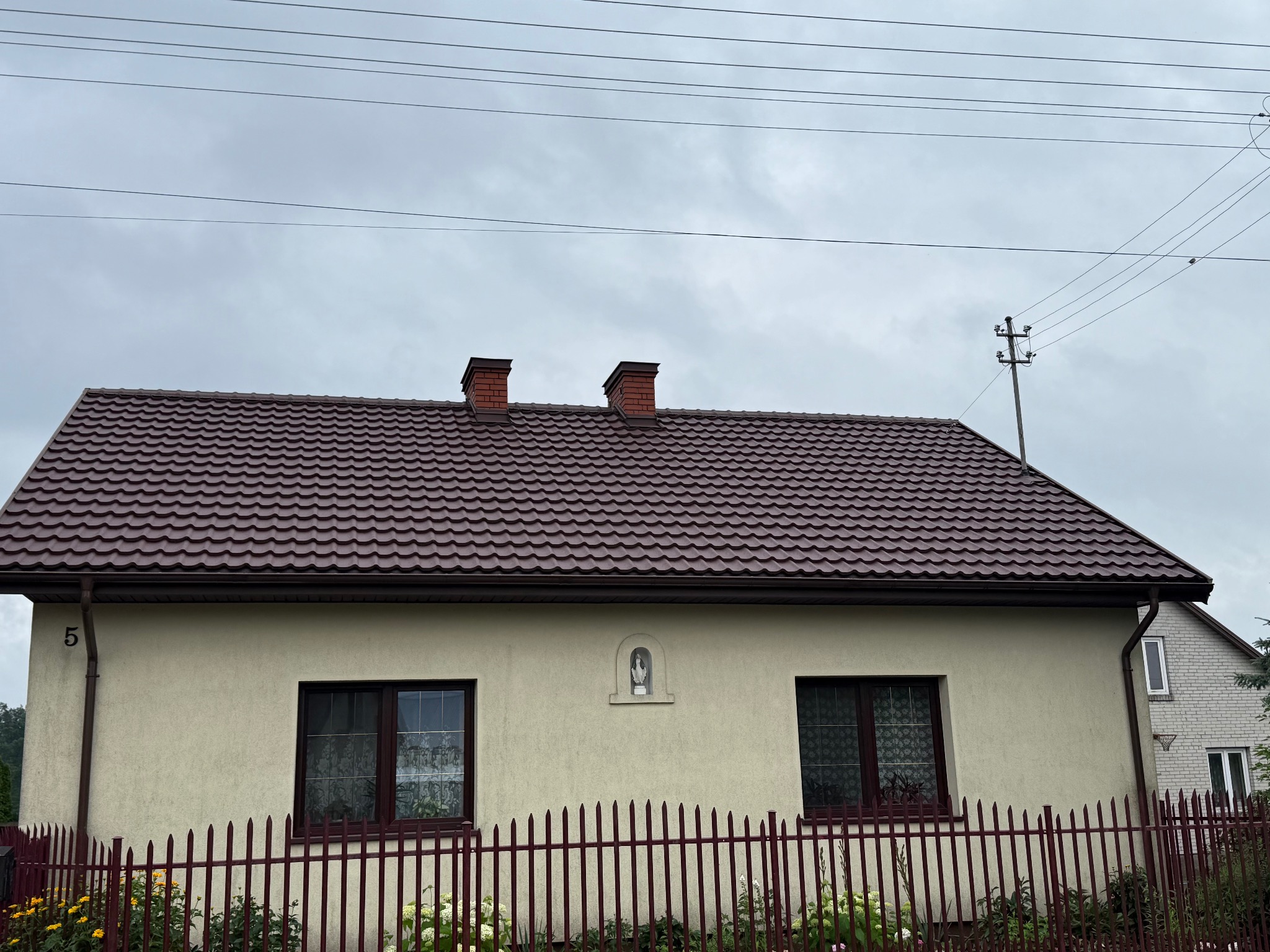 Dom jednorodzinny z brązowym dachem z dachówki, rynnami w tym samym kolorze i dwoma kominami. Widoczny numer domu '5' i ogrodzenie z czerwonych sztachet.