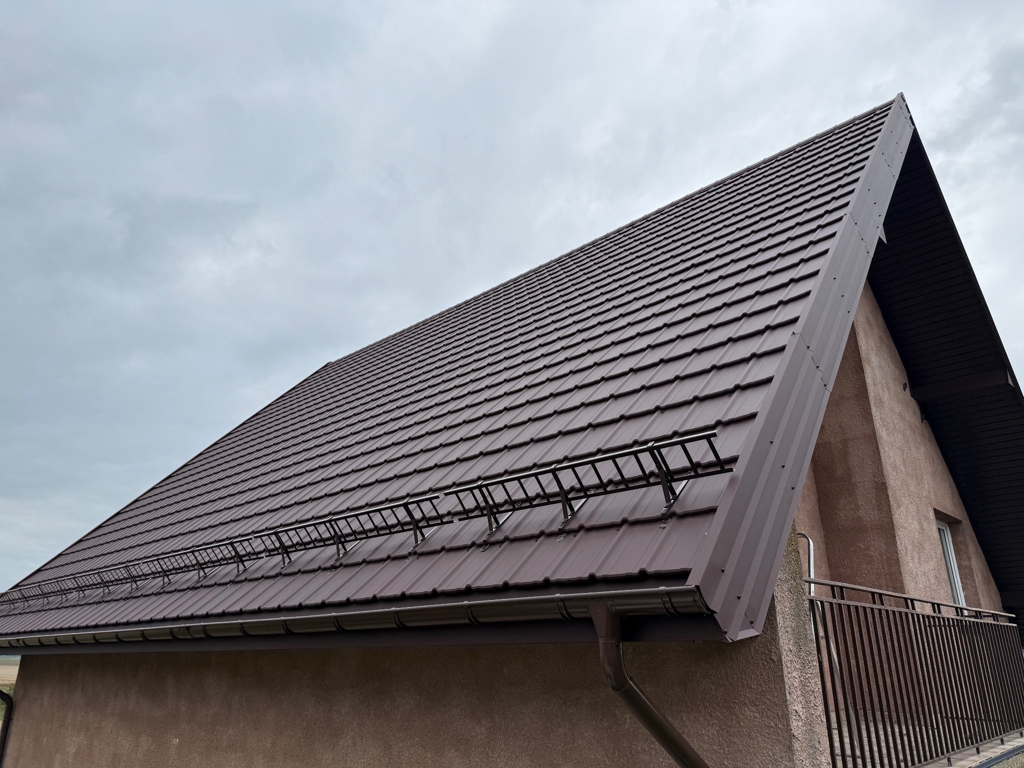 Brązowy dach z metalową konstrukcją przeciwśniegową i rynnami, widziany pod kątem na tle pochmurnego nieba. Widoczny fragment elewacji budynku.