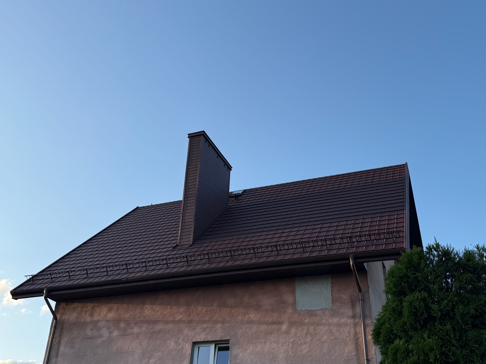 Ciemnobrązowy dach z kominem i rynnami na tle błękitnego nieba w Przasnyszu. Widok z dołu, ukazujący detale pokrycia i obróbki blacharskie.