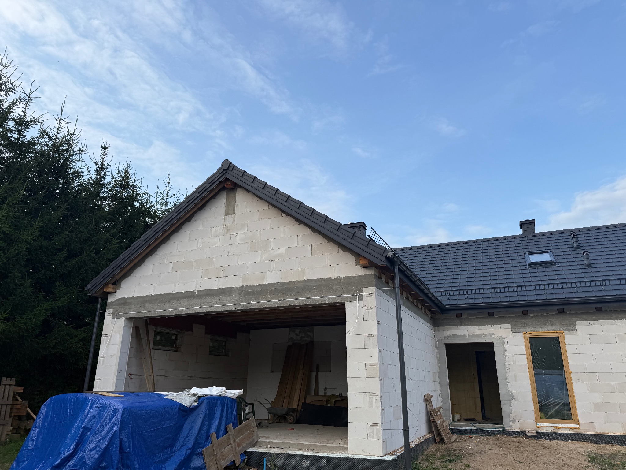 Budowa domu z białych bloczków, widoczny fragment dachu z czarną dachówką, okna wstawione, garaż otwarty, niebieska plandeka przykrywa materiały budowlane.