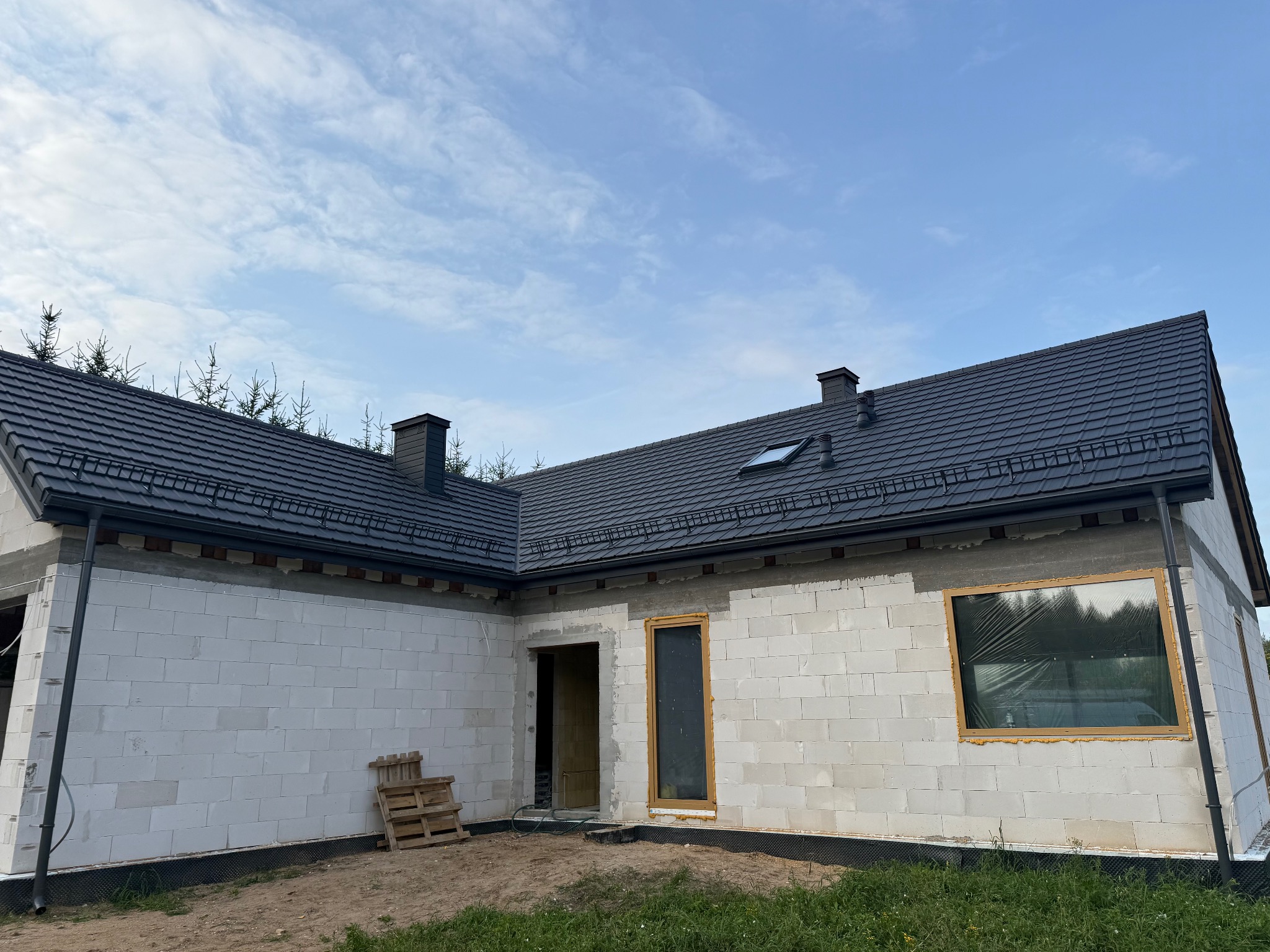 Dom jednorodzinny w trakcie budowy: ściany z białego betonu komórkowego, grafitowy dach z dachówki, widoczne rynny i kominy. Częściowo zamontowana stolarka okienna.