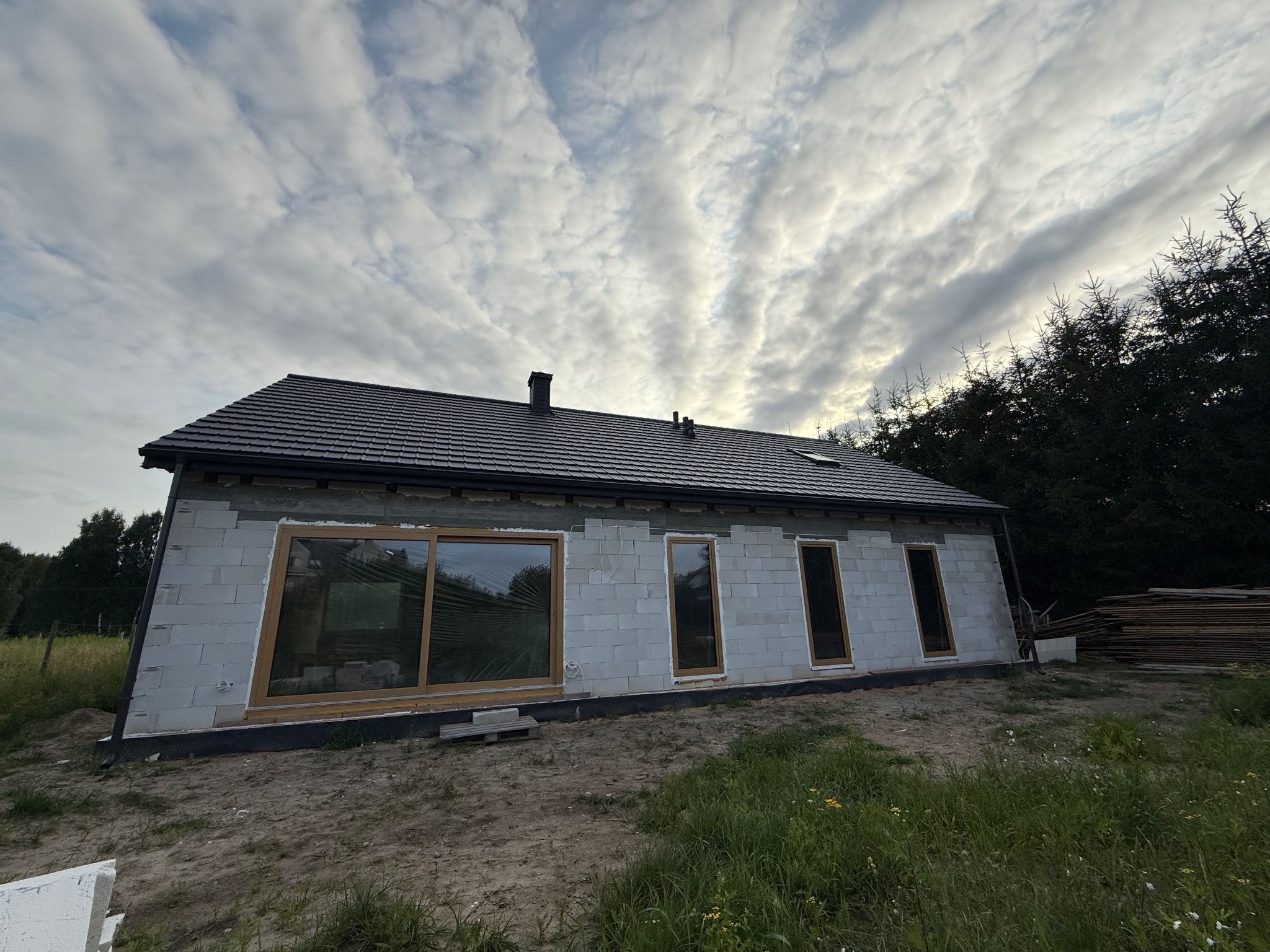 Dom w trakcie budowy z ciemną dachówką na skośnym dachu i oknami w drewnianych ramach. Widoczne bloczki betonowe ścian i elementy konstrukcyjne. Pochmurne niebo w tle.